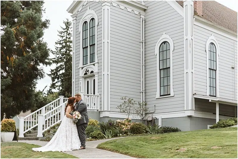 A bride and groom stand embracing and smiling in front of a light gray Snohomish church with tall arched windows, surrounded by green grass and trees on a cloudy day. Snohomish Wedding Photography