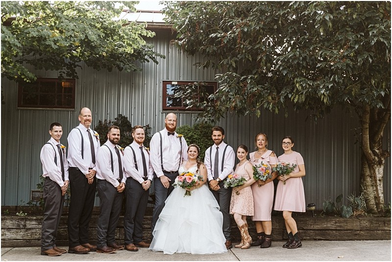 Premier snohomish & seattle wedding photographer | gsquared weddings photography a wedding party poses outdoors in front of a metal building at a snohomish seattle wedding The bride in a white gown is surrounded by smiling bridesmaids in pink and groomsmen with suspenders all holding bouquets and ready to celebrate the special day | serving snohomish seattle                   & beyond