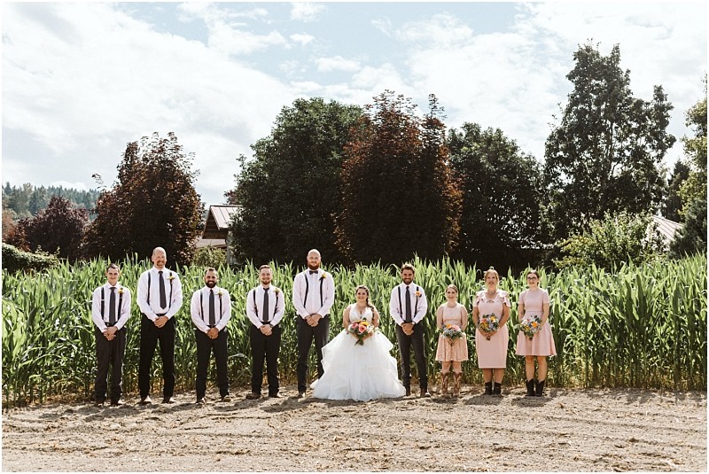 Premier snohomish & seattle wedding photographer | gsquared weddings photography a wedding party poses outdoors in front of tall green plants and trees at a beautiful snohomish seattle wedding The bride stands in the center in a white dress surrounded by bridesmaids in pink and groomsmen with suspenders | serving snohomish seattle                   & beyond