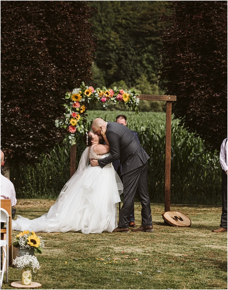 Premier snohomish & seattle wedding photographer | gsquared weddings photography a bride and groom share a kiss under a flower covered wooden arch during their snohomish outdoor wedding ceremony surrounded by lush greenery and guests seated on the lawn | serving snohomish seattle                   & beyond