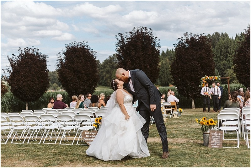 Premier snohomish & seattle wedding photographer | gsquared weddings photography a bride and groom share a kiss outdoors during their snohomish wedding ceremony surrounded by guests on white chairs and lush greenery with sunflowers and trees in the background | serving snohomish seattle                   & beyond
