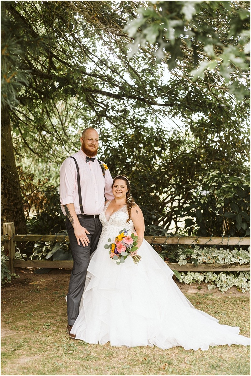 Premier snohomish & seattle wedding photographer | gsquared weddings photography a bride in a white gown holding a colorful bouquet stands beside a groom in dress pants suspenders and a light pink shirt Smiling outdoors they create unforgettable snohomish seattle wedding memories amid leafy trees and a wooden fence | serving snohomish seattle                   & beyond