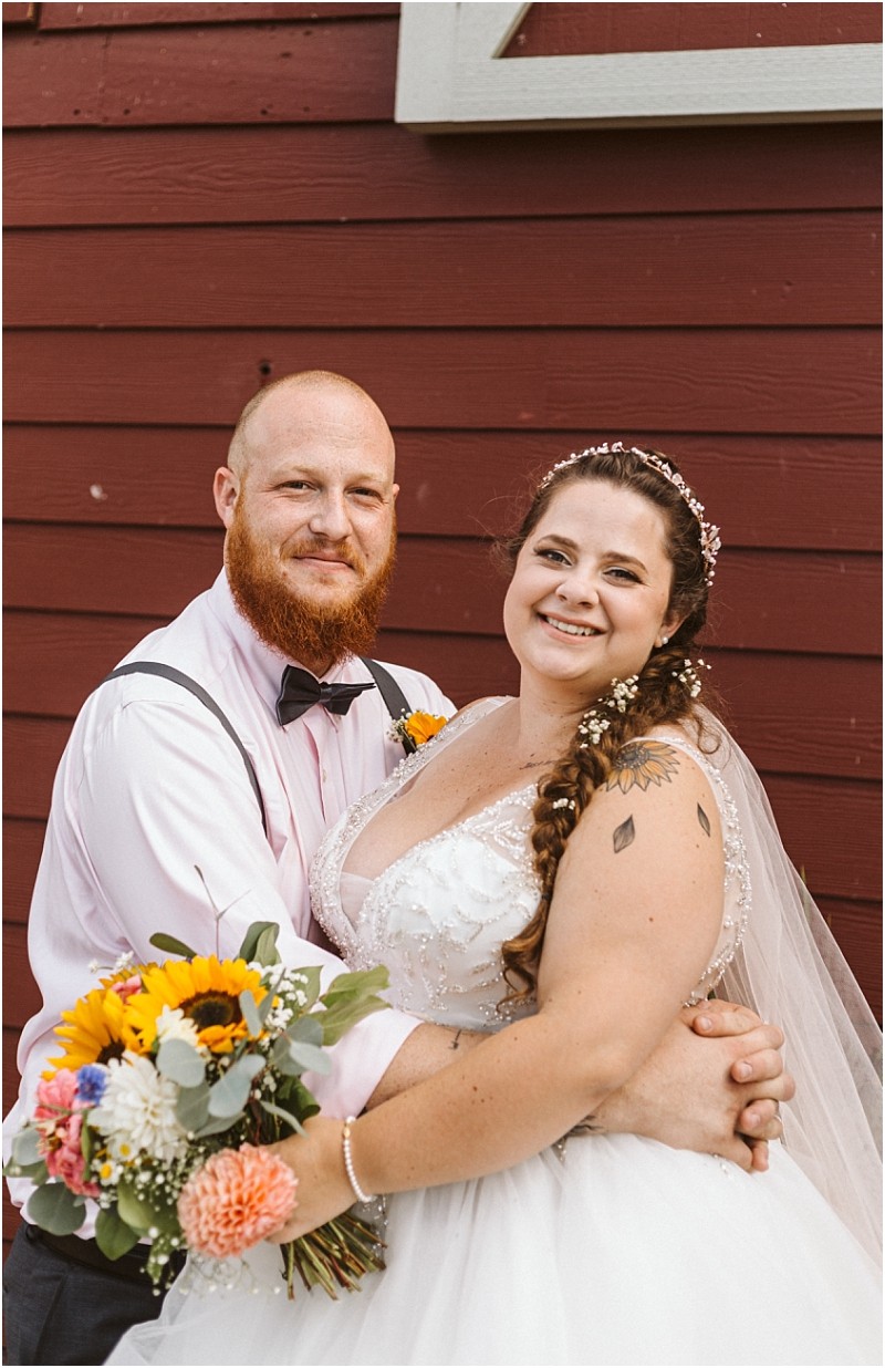 Premier snohomish & seattle wedding photographer | gsquared weddings photography a bride and groom stand smiling in front of a red wooden wall at their snohomish wedding The bride holds a colorful bouquet with sunflowers the groom wears a light pink shirt bow tie and suspenders | serving snohomish seattle                   & beyond