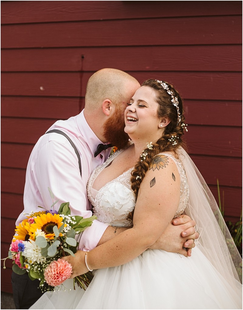 Premier snohomish & seattle wedding photographer | gsquared weddings photography a bride and groom embrace joyfully in front of a red wooden wall at their snohomish seattle wedding The groom kisses the brides head as she smiles brightly holding a colorful bouquet both dressed in elegant wedding attire | serving snohomish seattle                   & beyond