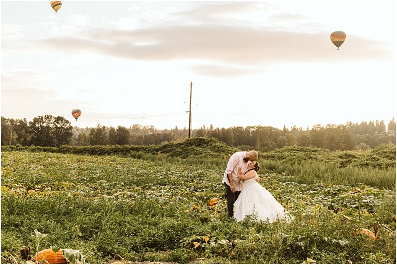 Premier snohomish & seattle wedding photographer | gsquared weddings photography a couple in wedding attire embraces and kisses in a lush green field with pumpkins under a bright sky with hot air balloons floating in the distancecapturing the magic of a snohomish seattle wedding engagement | serving snohomish seattle                   & beyond