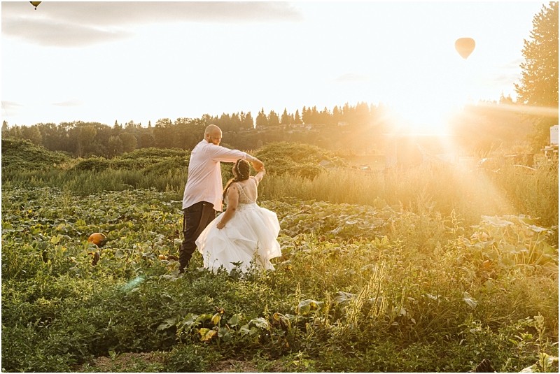 Premier snohomish & seattle wedding photographer | gsquared weddings photography a couple dances in a lush green field at sunset during their snohomish seattle wedding engagement with the sun shining brightly behind them and a hot air balloon floating in the sky The woman wears a white dress and the man wears a light shirt and dark pants | serving snohomish seattle                   & beyond