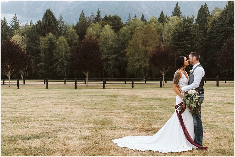 Premier snohomish & seattle wedding photographer | gsquared weddings photography a bride and groom stand close together kissing and holding a bouquet in a grassy snohomish field with trees and mountains in the background The bride wears a white gown with a long train capturing the romance of their seattle wedding engagement | serving snohomish seattle                   & beyond