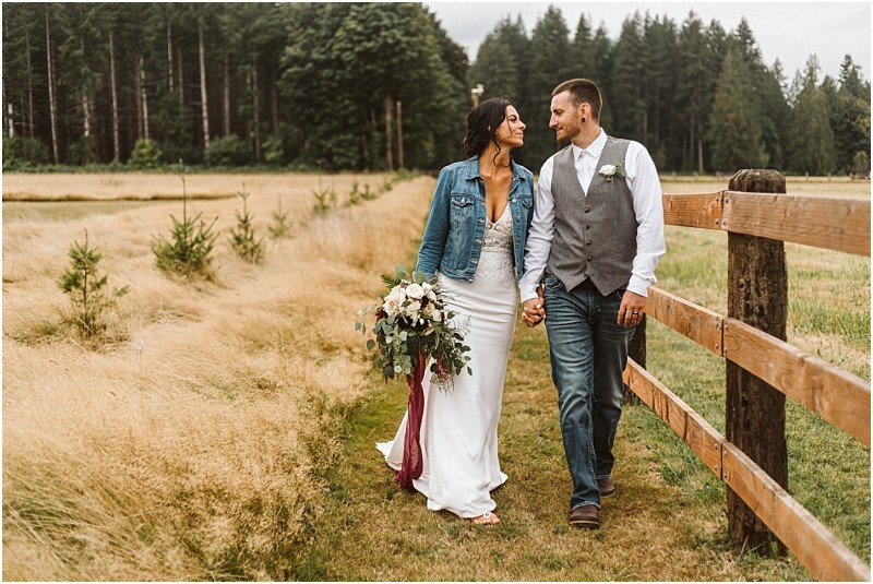 Premier snohomish & seattle wedding photographer | gsquared weddings photography a bride and groom walk hand in hand along a wooden fence in a grassy field at their snohomish wedding The bride wears a white dress and denim jacket holding her bouquet while the groom wears jeans a vest and white shirt Trees frame the scene | serving snohomish seattle                   & beyond