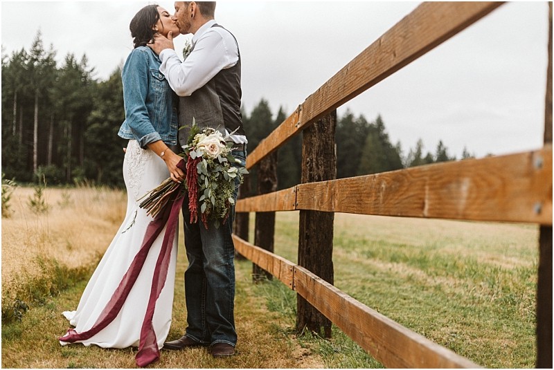 Premier snohomish & seattle wedding photographer | gsquared weddings photography a bride in a white dress and denim jacket kisses her groom in a vest and jeans beside a wooden fence in a grassy field holding a bouquet with burgundy ribbons Forest trees hint at the rustic charm of a snohomish seattle wedding | serving snohomish seattle                   & beyond