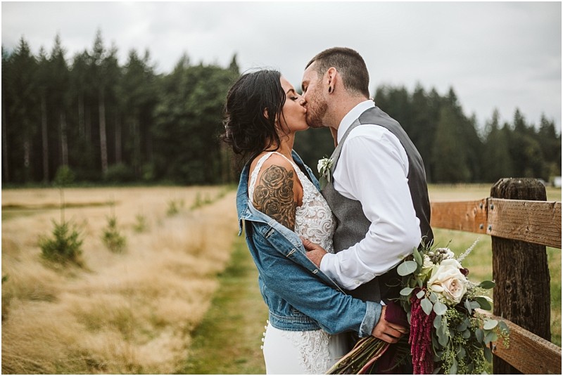 Premier snohomish & seattle wedding photographer | gsquared weddings photography a bride in a white dress and denim jacket kisses her groom in a vest and tie beside a wooden fence holding a bouquet during their snohomish wedding with a grassy field and trees in the background | serving snohomish seattle                   & beyond
