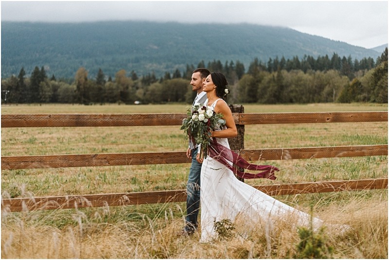 Premier snohomish & seattle wedding photographer | gsquared weddings photography a bride and groom walk side by side along a wooden fence in a grassy field near snohomish with mountains and trees in the background The bride holds a bouquet and wears a flowing white dress the groom is in jeans and a vest | serving snohomish seattle                   & beyond