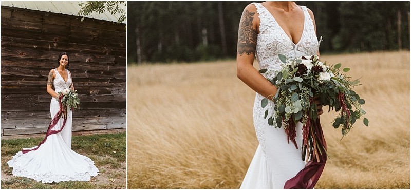 Premier snohomish & seattle wedding photographer | gsquared weddings photography a bride in a fitted white lace dress with a long train stands outdoors at a rustic snohomish venue holding a large bouquet of greenery and white flowers with a burgundy ribbon She smiles beside a wooden wall her arm tattoos visibleperfect for any seattle wedding inspiration | serving snohomish seattle                   & beyond