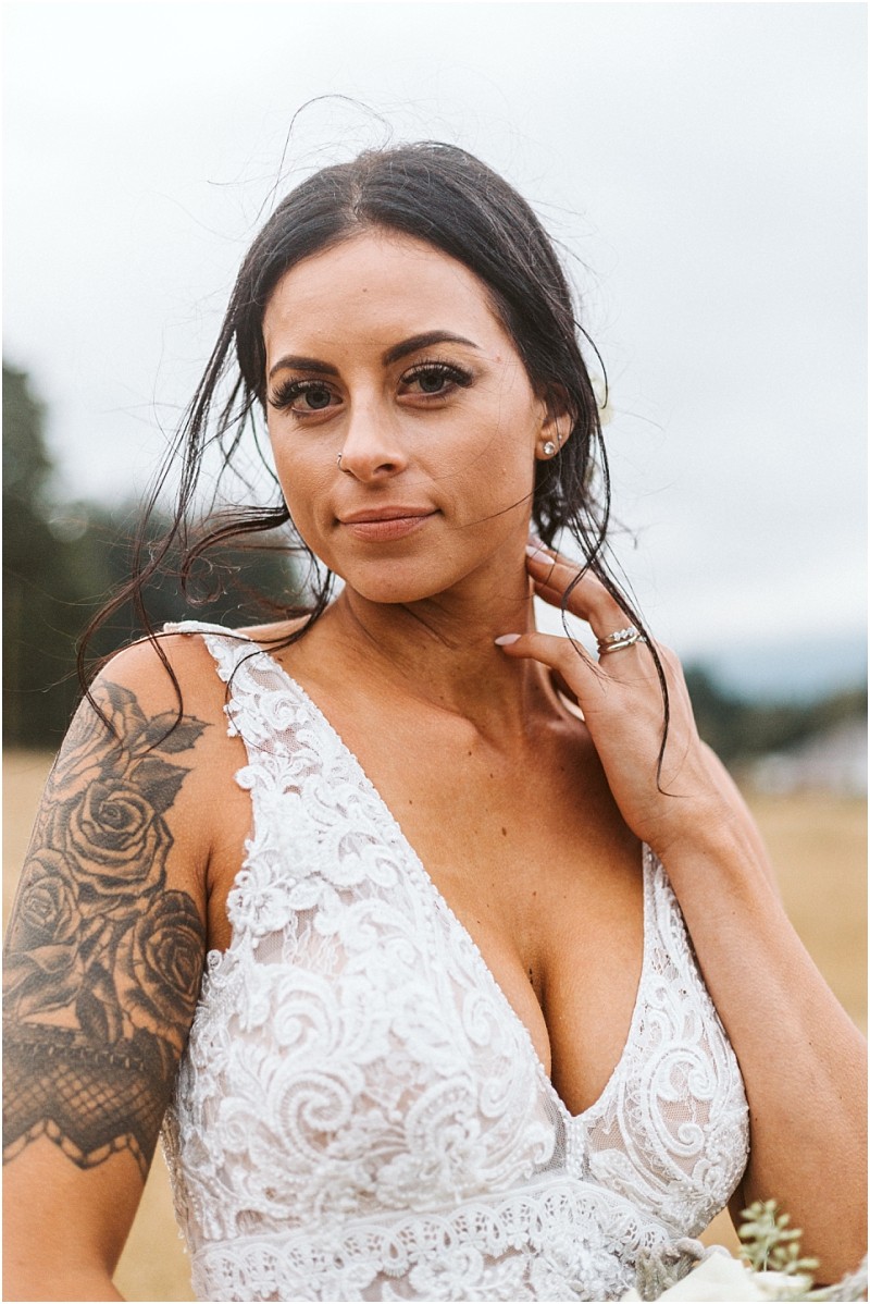 Premier snohomish & seattle wedding photographer | gsquared weddings photography a woman with long dark hair in a white lace dress stands outdoors near snohomish touching her neck She has a floral tattoo on her upper arm and wears a ring with a calm expression and a blurred natural background | serving snohomish seattle                   & beyond