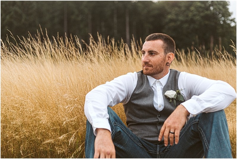Premier snohomish & seattle wedding photographer | gsquared weddings photography a man with short hair and a beard sits in tall golden grass at a snohomish seattle wedding engagement He wears a white shirt gray vest jeans and boutonniere looking thoughtfully into the distance with trees in the background | serving snohomish seattle                   & beyond