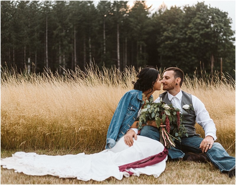 Premier snohomish & seattle wedding photographer | gsquared weddings photography a bride in a white dress and denim jacket sits in tall grass holding a bouquet and kisses her groom in a vest and jeans during their snohomish wedding Trees frame the scene under a cloudy seattle sky | serving snohomish seattle                   & beyond
