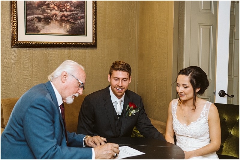 Premier snohomish & seattle wedding photographer | gsquared weddings photography a man in a blue suit signs a document at a table with a smiling bride in a white dress and groom in a dark suit both seated and holding hands during their snohomish seattle wedding A framed painting hangs on the wall behind them | serving snohomish seattle                   & beyond