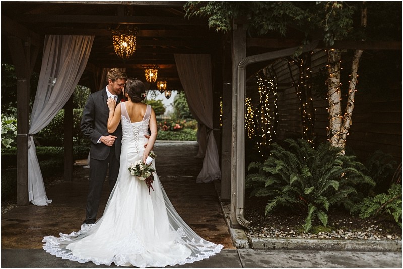 Premier snohomish & seattle wedding photographer | gsquared weddings photography a bride and groom stand close together under a covered outdoor walkway decorated with hanging lights and white drapes at their snohomish seattle wedding The bride holds a bouquet with fairy lights wrapped around a nearby tree adding to the romance | serving snohomish seattle                   & beyond