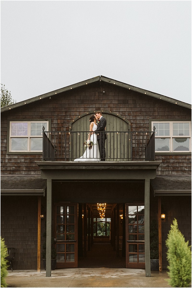 Premier snohomish & seattle wedding photographer | gsquared weddings photography a bride and groom stand close together on a balcony outside a rustic dark wood building in snohomish sharing a kiss in their wedding attire String lights decorate the upper edge capturing a romantic seattle wedding moment | serving snohomish seattle                   & beyond