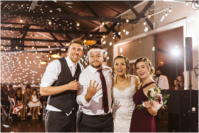 Premier snohomish & seattle wedding photographer | gsquared weddings photography four people pose and smile at a snohomish wedding reception the groom with a garter on his head the bride in white a bridesmaid in burgundy holding flowers and a groomsman String lights and guests highlight this seattle celebration | serving snohomish seattle                   & beyond