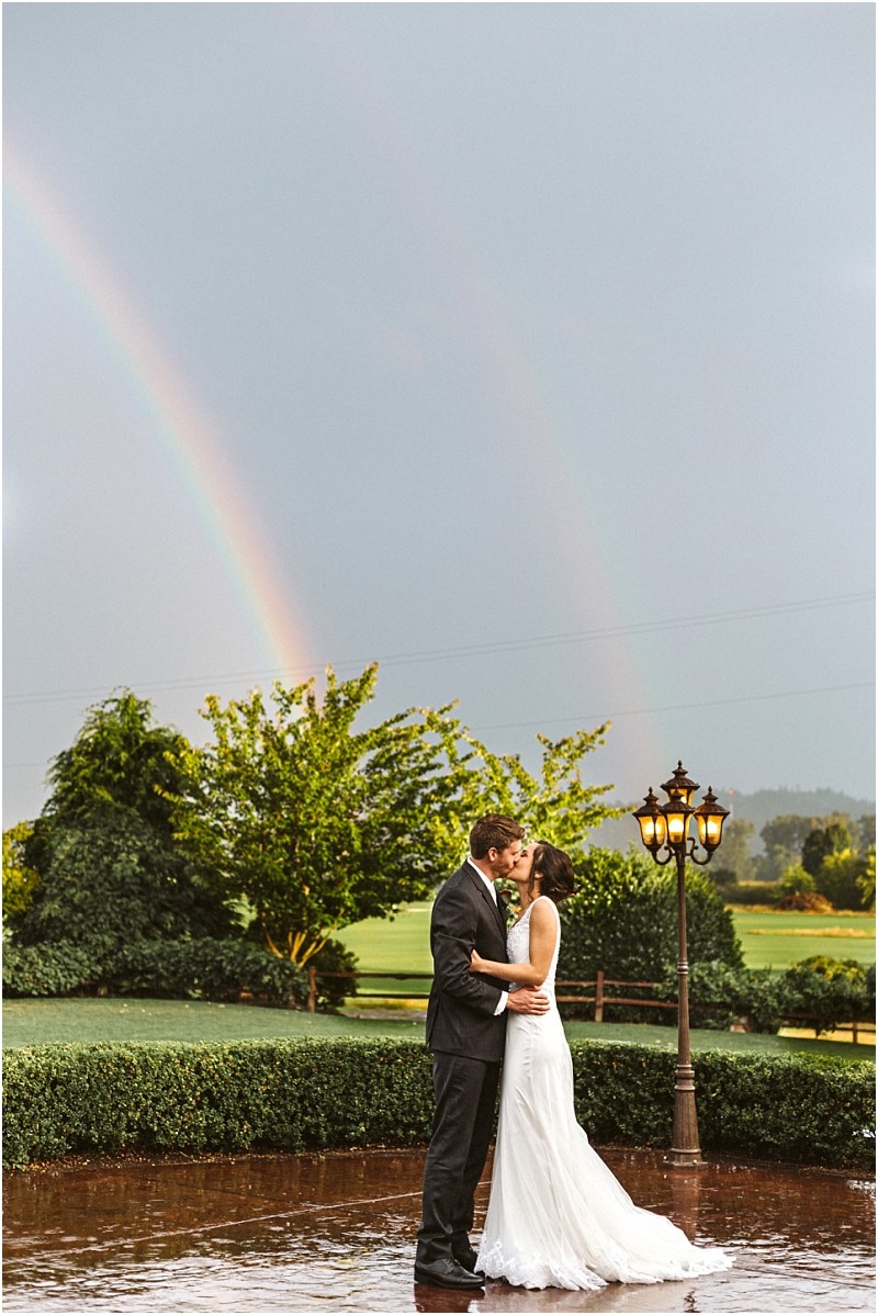 Premier snohomish & seattle wedding photographer | gsquared weddings photography a bride and groom embrace and kiss outdoors by a vintage lamp post lush green trees and a vivid double rainbow in the cloudy sky behind themcapturing a magical snohomish seattle wedding moment reflected on the wet ground | serving snohomish seattle                   & beyond