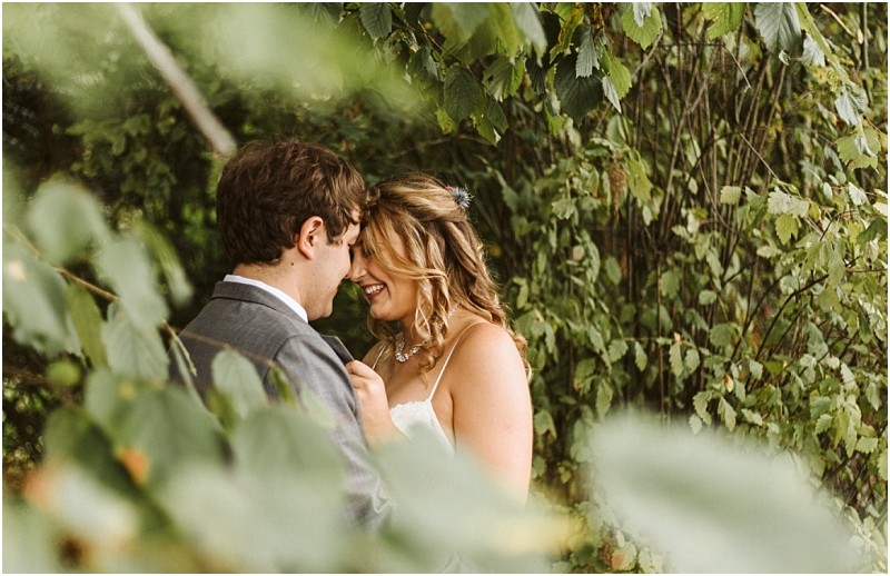 Premier snohomish & seattle wedding photographer | gsquared weddings photography a bride and groom stand close together smiling and touching foreheads partially hidden by green leafy branches in a beautiful snohomish outdoor settingcapturing the romance of a seattle wedding engagement | serving snohomish seattle                   & beyond