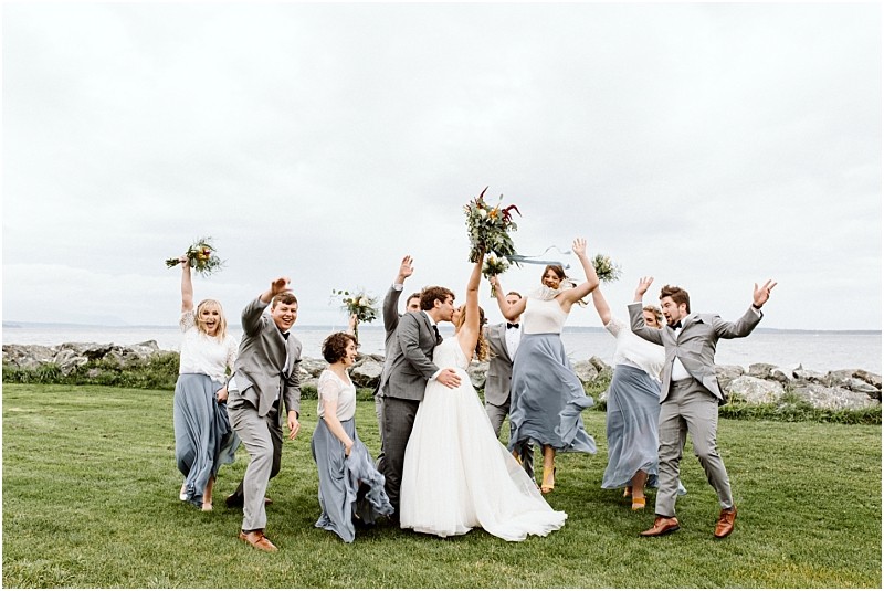 Premier snohomish & seattle wedding photographer | gsquared weddings photography a wedding party poses joyfully on a grassy lawn by the water at a stunning snohomish seattle wedding The bride and groom stand center surrounded by bridesmaids in blue dresses and groomsmen in gray suits all celebrating with lifted bouquets | serving snohomish seattle                   & beyond