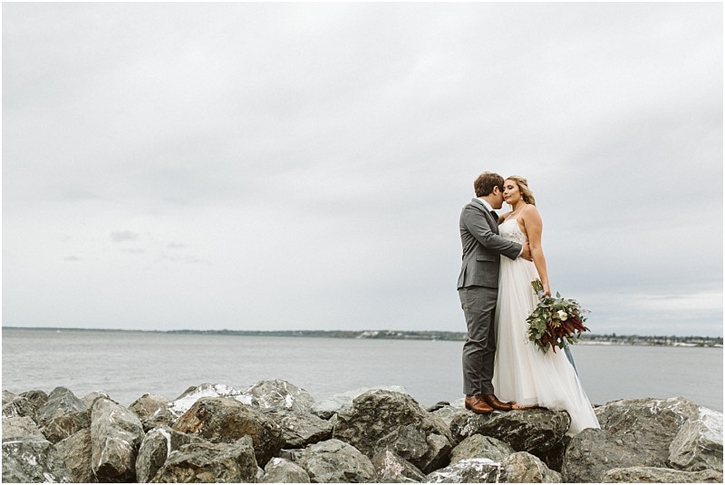 Premier snohomish & seattle wedding photographer | gsquared weddings photography a bride and groom stand on a rocky shore by the water near seattle embracing and kissing under a cloudy sky The bride holds a bouquet and wears a white dress while the groom in a gray suit shares this romantic snohomish wedding moment | serving snohomish seattle                   & beyond