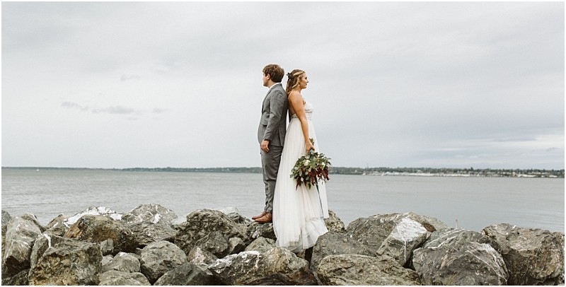 Premier snohomish & seattle wedding photographer | gsquared weddings photography a bride and groom stand back to back on large rocks by the water facing opposite directions The overcast sky and distant shoreline set a serene moodperfect for a snohomish seattle wedding or engagement The bride holds a bouquet of flowers | serving snohomish seattle                   & beyond