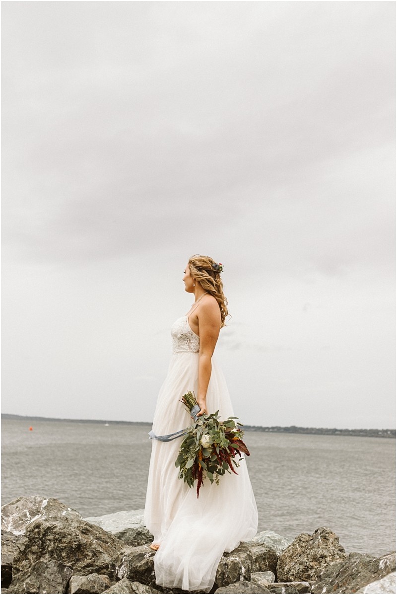 Premier snohomish & seattle wedding photographer | gsquared weddings photography a bride in a white gown stands on rocky ground by a body of water near snohomish holding a bouquet of greenery and flowers gazing into the distance under a cloudy skycapturing the magic of a seattle wedding engagement | serving snohomish seattle                   & beyond