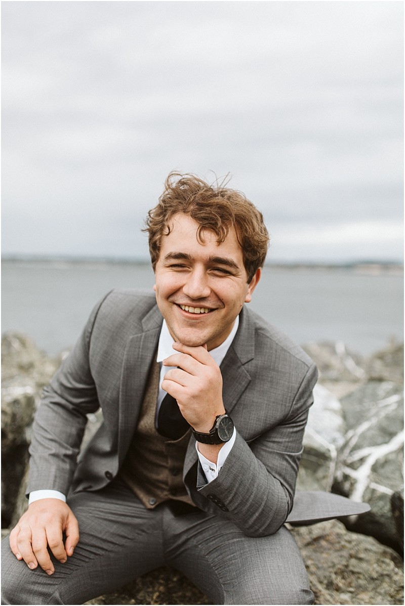 Premier snohomish & seattle wedding photographer | gsquared weddings photography a young man in a gray suit sits on rocks by the water smiling warmly at the camera with his hand resting on his chin The overcast sky and blurred shoreline create a serene backdrop for this snohomish seattle engagement moment | serving snohomish seattle                   & beyond