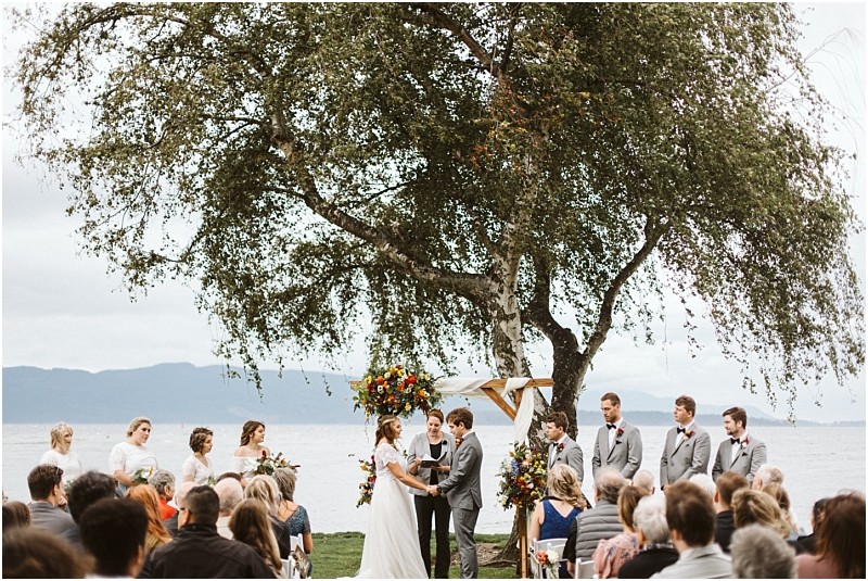 Premier snohomish & seattle wedding photographer | gsquared weddings photography a bride and groom stand under a floral arch by a large tree exchanging vows at their snohomish outdoor wedding ceremony by the water surrounded by their wedding party and seated guests | serving snohomish seattle                   & beyond