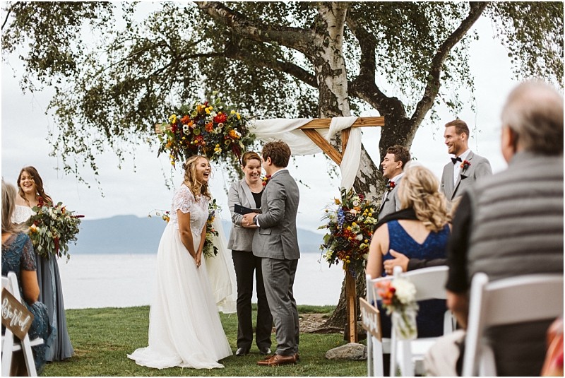 Premier snohomish & seattle wedding photographer | gsquared weddings photography a bride and groom stand smiling under a decorated wooden arch by a lake during an outdoor snohomish wedding ceremony surrounded by their wedding party and seated guests | serving snohomish seattle                   & beyond