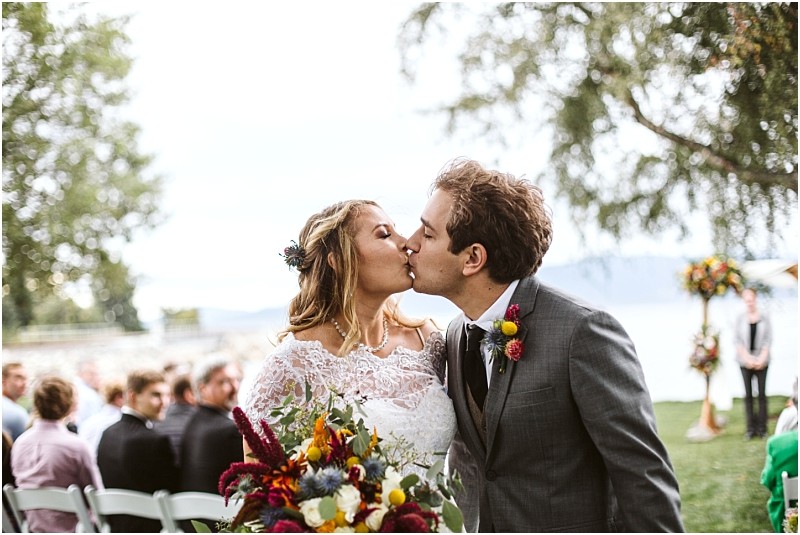 Premier snohomish & seattle wedding photographer | gsquared weddings photography a newlywed couple shares a kiss outdoors after their snohomish wedding ceremony holding a vibrant bouquet with guests seated behind them and a lake and floral arch in the background | serving snohomish seattle                   & beyond