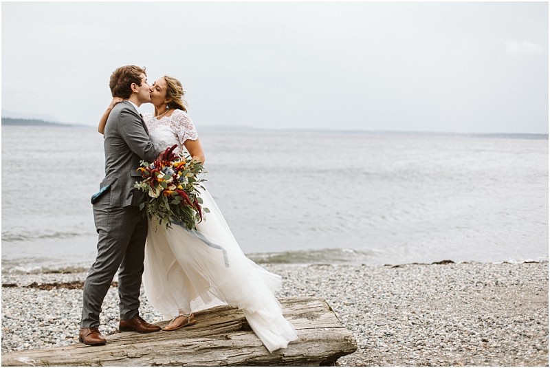 Premier snohomish & seattle wedding photographer | gsquared weddings photography a bride and groom kiss on a rocky beach near snohomish standing on driftwood The bride holds a colorful bouquet in her white dress while the groom wears a gray suit The sea and cloudy sky set a romantic seattle wedding scene | serving snohomish seattle                   & beyond