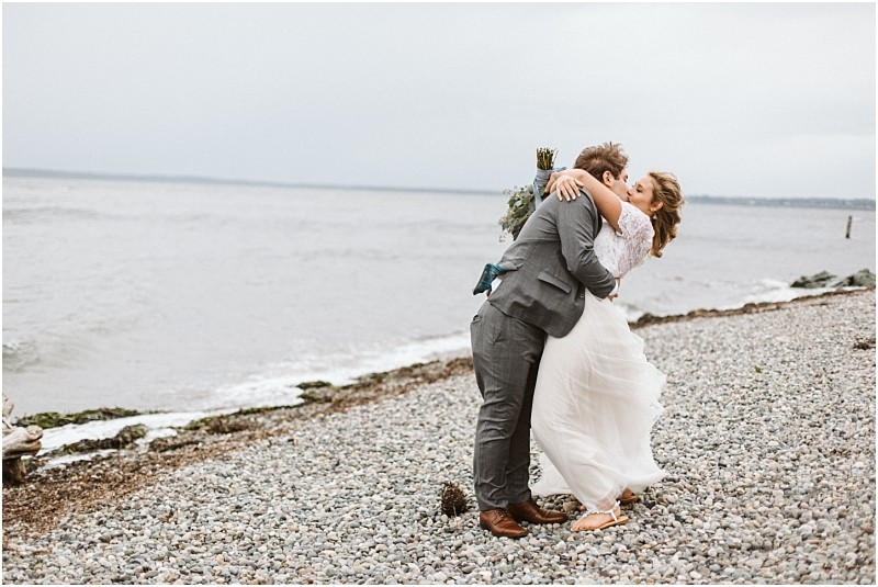 Premier snohomish & seattle wedding photographer | gsquared weddings photography a newlywed couple embraces and shares a kiss on a rocky snohomish beach with the ocean and cloudy sky in the background The bride wears a flowing white dress and the groom is in a gray suit capturing their romantic seattle wedding | serving snohomish seattle                   & beyond