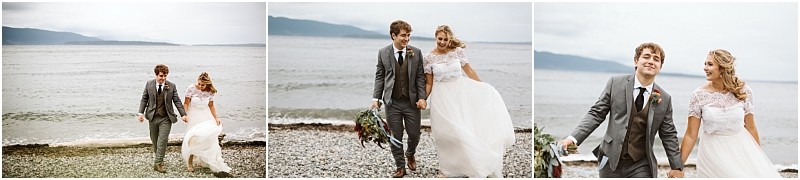 Premier snohomish & seattle wedding photographer | gsquared weddings photography a bride and groom walk hand in hand along a rocky beach near snohomish smiling and laughing together The bride wears a white dress and the groom wears a gray suit with calm water and mountains framing their seattle wedding day | serving snohomish seattle                   & beyond