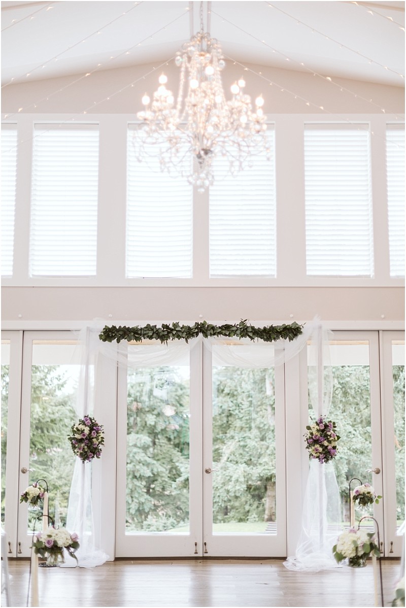 Premier snohomish & seattle wedding photographer | gsquared weddings photography elegant indoor snohomish wedding ceremony setup with tall windows a sparkling chandelier white drapery green garland and purple floral arrangements framing the glass doors with trees visible outside | serving snohomish seattle                   & beyond