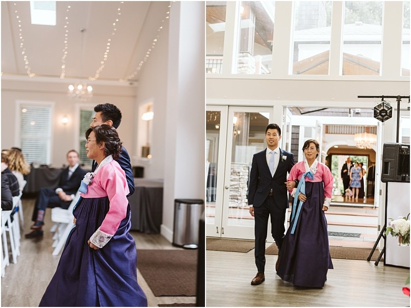 Premier snohomish & seattle wedding photographer | gsquared weddings photography at a snohomish seattle wedding a woman in a pink and navy hanbok walks beside a man in a suit at an indoor event possibly an engagement with guests seated and string lights glowing above | serving snohomish seattle                   & beyond