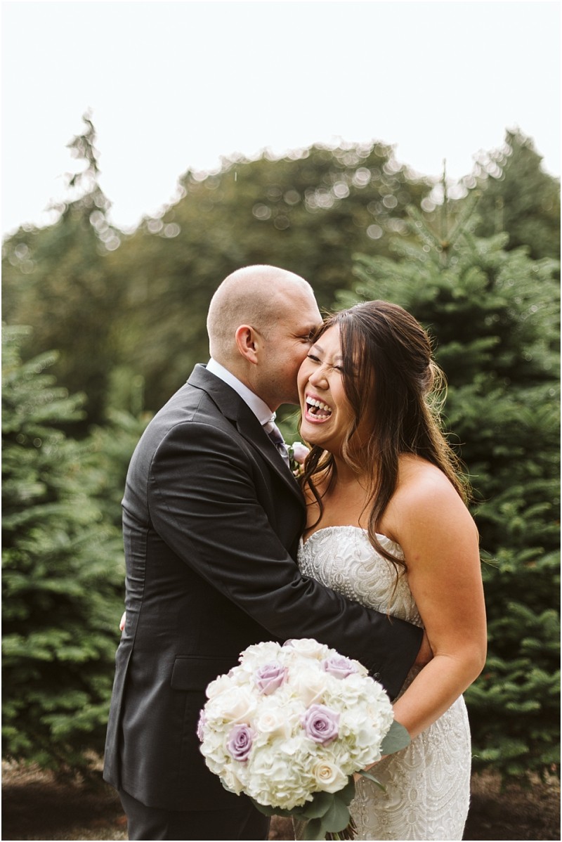 Premier snohomish & seattle wedding photographer | gsquared weddings photography a groom in a black suit kisses his smiling bride holding a bouquet of white and purple flowers They stand close together outdoors surrounded by green pine trees at their snohomish seattle wedding | serving snohomish seattle                   & beyond