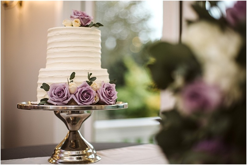 Premier snohomish & seattle wedding photographer | gsquared weddings photography a two tiered white cake decorated with purple and white roses sits on a silver cake standa perfect centerpiece for a snohomish or seattle wedding The softly blurred background reveals lush greenery through a window | serving snohomish seattle                   & beyond