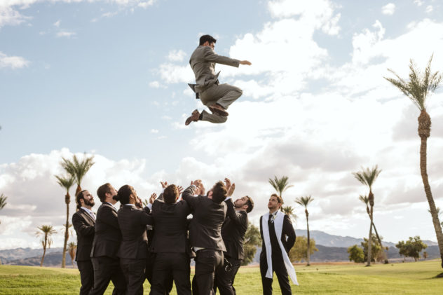 Premier snohomish & seattle wedding photographer | gsquared weddings photography a group of men in suits toss another man also in a suit high into the air outdoors on a grassy field at a snohomish seattle wedding with palm trees and mountains in the background under a partly cloudy sky | serving snohomish seattle                   & beyond