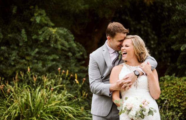 Premier snohomish & seattle wedding photographer | gsquared weddings photography a happy bride and groom stand outside smiling and laughing together during their snohomish seattle wedding The groom hugs the bride from behind as she holds a bouquet of flowers surrounded by lush green foliage | serving snohomish seattle                   & beyond