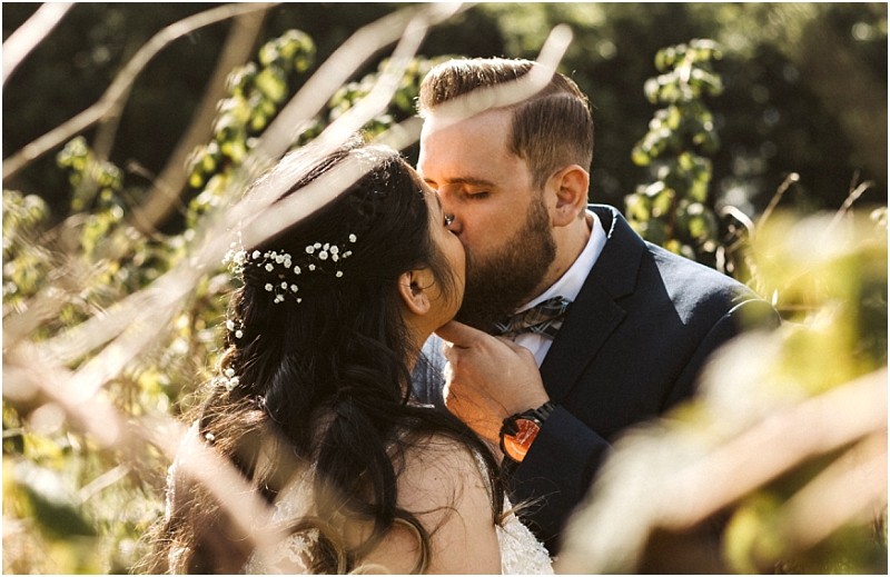 Premier Snohomish & Seattle Wedding Photographer | GSquared Weddings Photography A bride and groom share a kiss outdoors at their Snohomish Seattle wedding, surrounded by greenery. The bride’s dark hair is adorned with small white flowers, and the groom in a navy suit gently holds her chin as sunlight filters through branches.                                   | Serving Snohomish, Seattle ,                  & Beyond