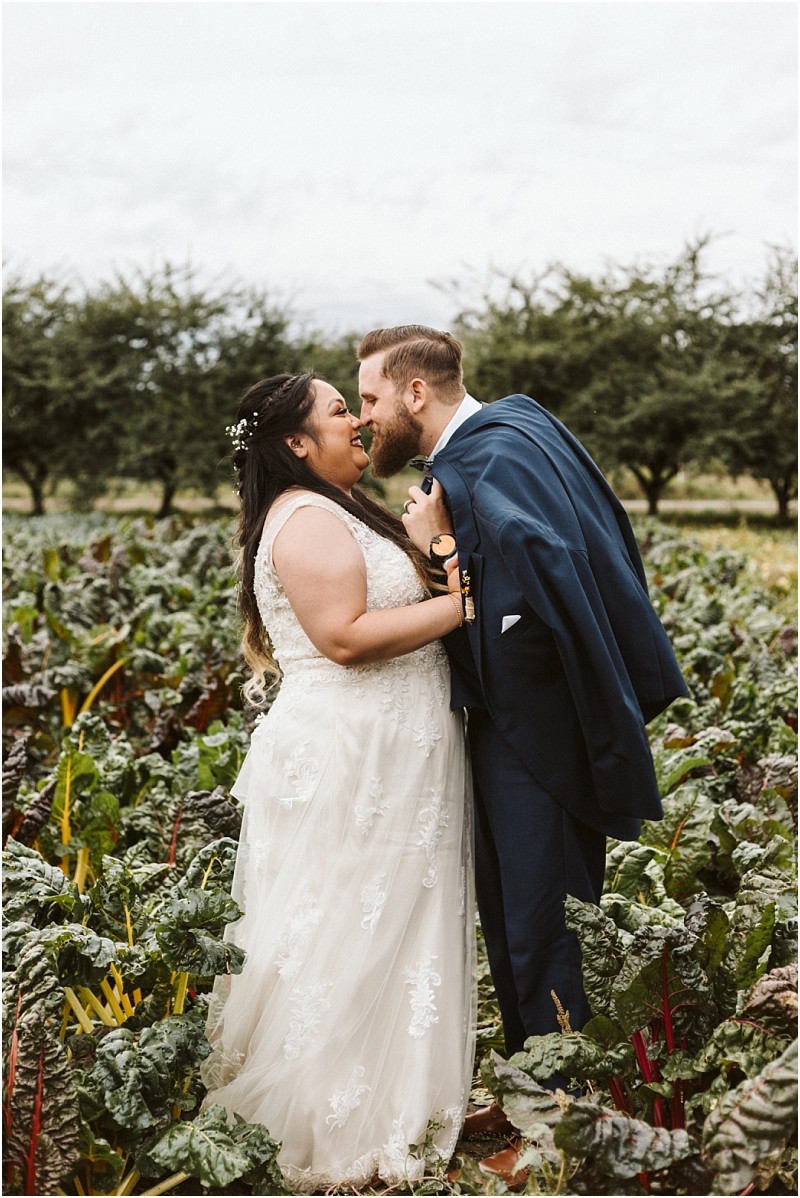 Premier snohomish & seattle wedding photographer | gsquared weddings photography a bride and groom stand closely together in a lush snohomish vegetable garden smiling and about to kiss The bride wears a white lace gown and the groom in a navy suit drapes his jacket over his shoulder Trees frame this seattle wedding moment | serving snohomish seattle                   & beyond