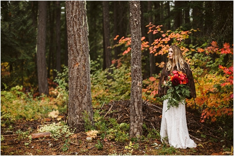 Premier snohomish & seattle wedding photographer | gsquared weddings photography a woman in a white dress and brown jacket stands in a forest near snohomish holding a bouquet of red and orange flowers Vibrant autumn leaves surround her with tall trees in the backgroundperfect for a seattle wedding engagement shoot | serving snohomish seattle                   & beyond