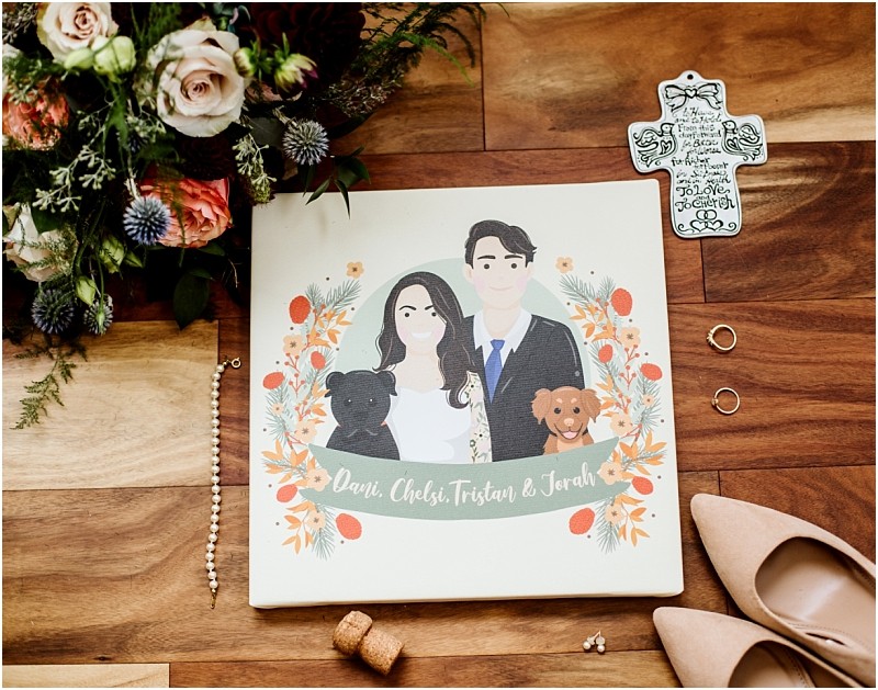 Premier snohomish & seattle wedding photographer | gsquared weddings photography a snohomish seattle wedding flat lay features a floral bouquet an illustrated portrait of the engaged couple with their two dogs wedding rings a decorative cross beige heels and a cork on a wooden floor The portrait reads dani chels tristan & jonah | serving snohomish seattle                   & beyond