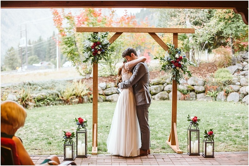 Premier snohomish & seattle wedding photographer | gsquared weddings photography a bride and groom embrace under a wooden wedding arch decorated with flowers and lanterns outdoors with greenery and autumn foliage in the background at a snohomish seattle wedding A seated guest is partially visible in the foreground | serving snohomish seattle                   & beyond