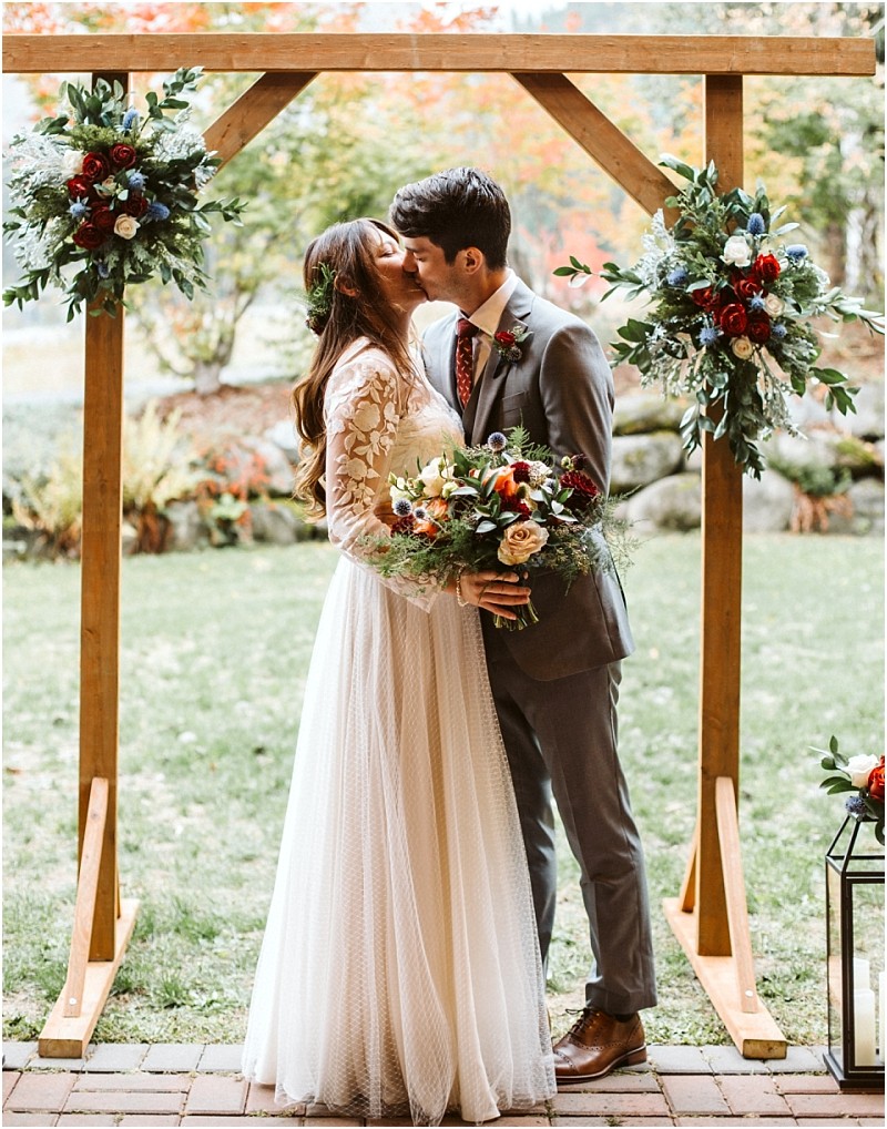 Premier snohomish & seattle wedding photographer | gsquared weddings photography a bride and groom kiss under a wooden arch decorated with flowers in a picturesque snohomish outdoor setting The bride holds a bouquet in her lace dress while the groom wears a gray suit and patterned tie | serving snohomish seattle                   & beyond