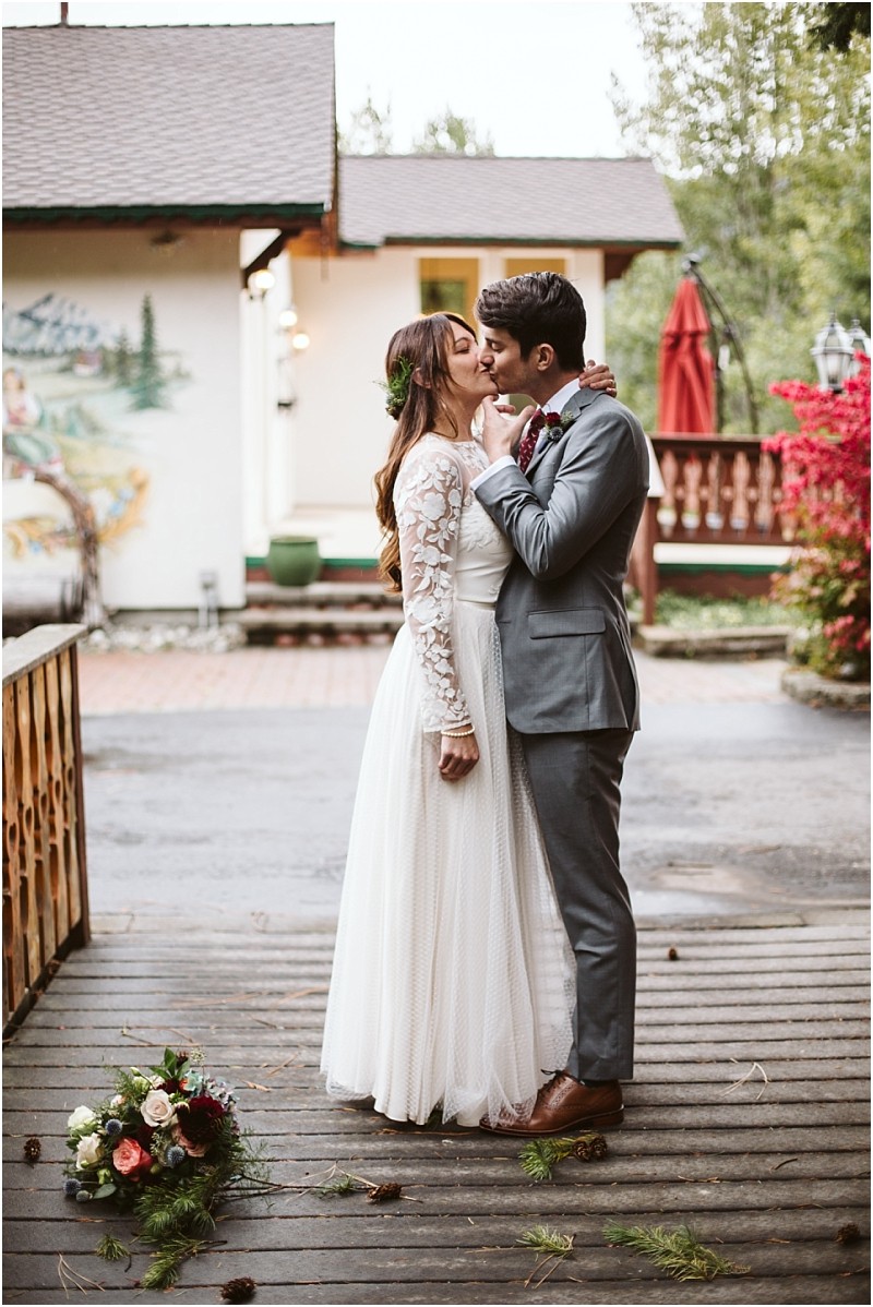 A bride in a white lace dress and a groom in a gray suit share a kiss outdoors on a wooden walkway in snohomish with a bouquet on the ground and a rustic building in the background Snohomish wedding photography