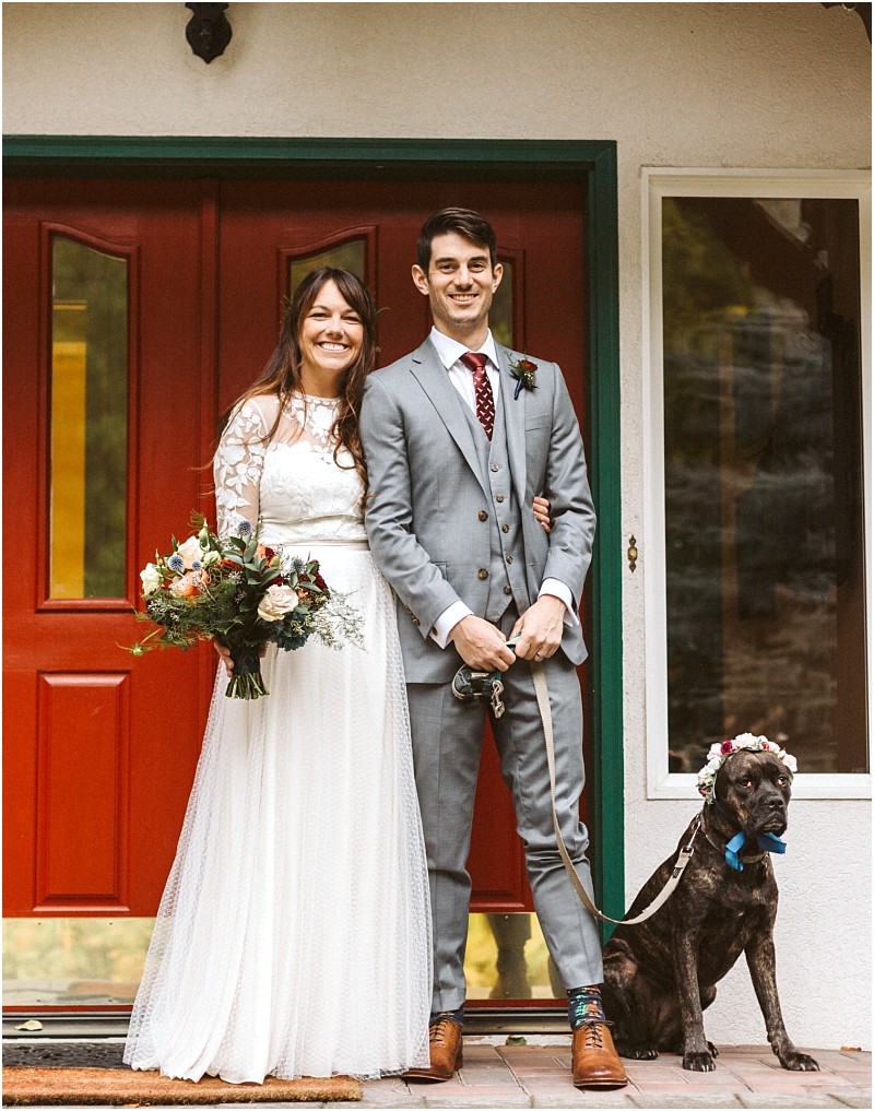 Premier snohomish & seattle wedding photographer | gsquared weddings photography a smiling bride in a white lace dress stands beside a groom in a gray suit holding a leash Their dog wearing a flower crown sits near them in front of a red door at their dreamy snohomish seattle wedding | serving snohomish seattle                   & beyond